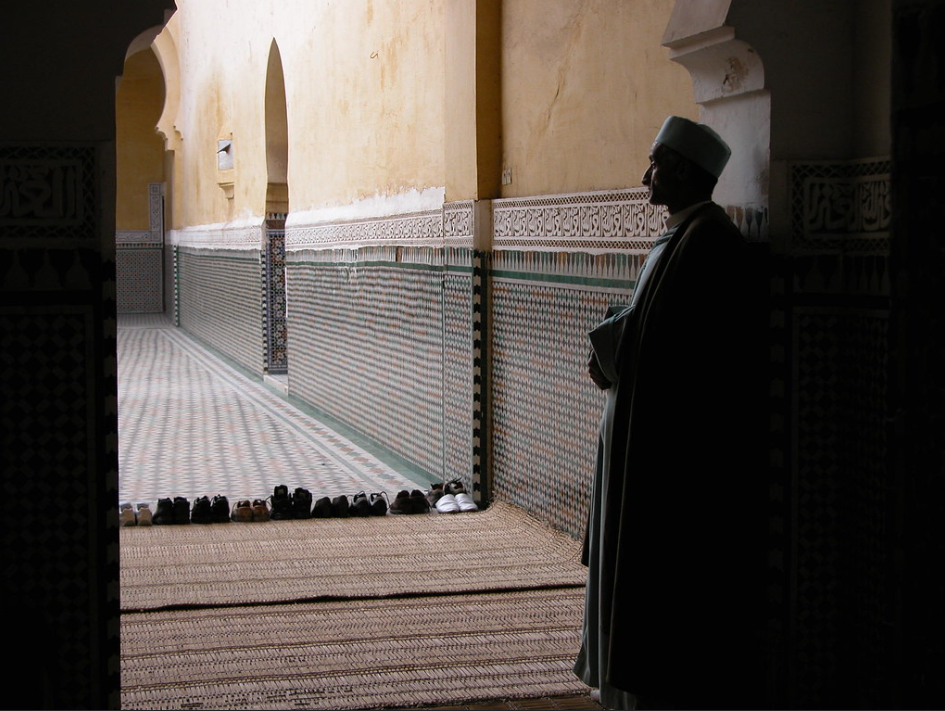 Muslim man standing against the wall with hands folded, solemnly looking on while watching others in prayer.